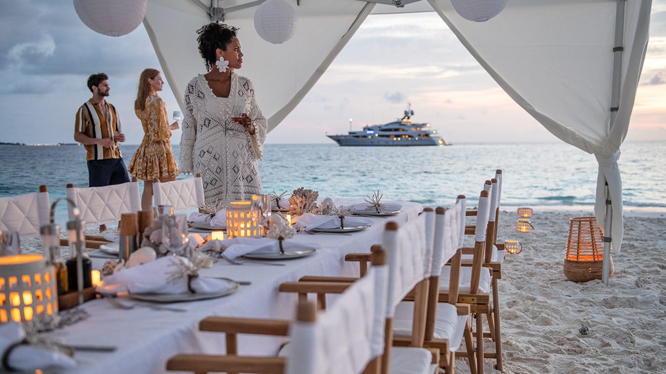 Beachside dinner setup with a view of a yacht charter at sunset, part of the exclusive Edmiston luxury experience.
