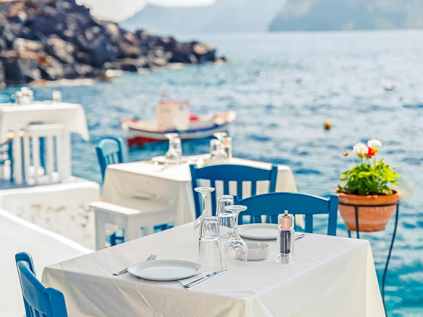 A seaside Greek restaurant has tables set, awaiting guests for a private luxury escape on a Mediterranean superyacht charter.