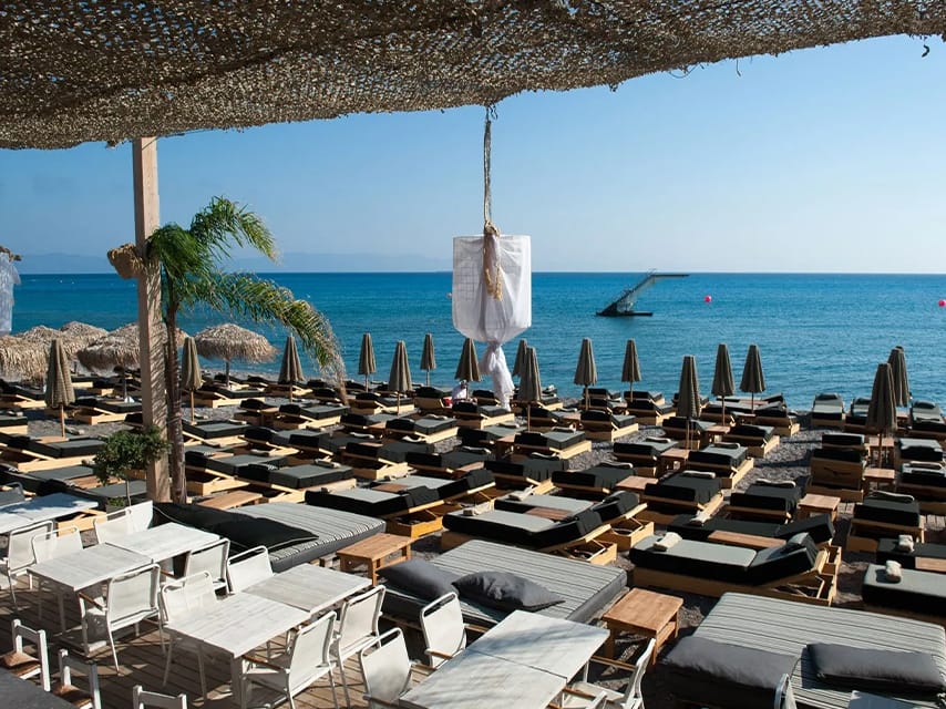 Dining tables and sunbeds overlook the sea at Ronda Beach Bar, Rhodes, with stunning views and gourmet food, a must-visit on your private yacht charter in Greece.