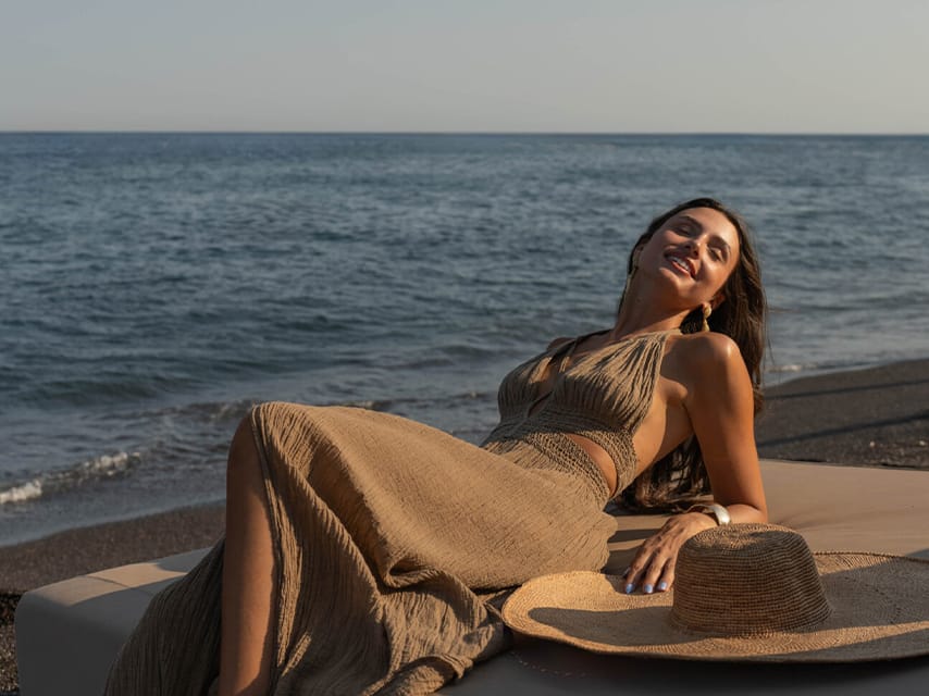 At the best beach club in Santorini, a woman in a taupe boho dress lounges on a beachside bed, as part of her luxury yacht charter in the Greek Islands.