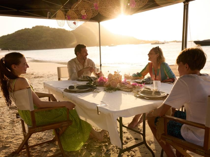 A family of four enjoys a private beach dinner at sunset during a sailing yacht charter Mediterranean voyage in Greece.