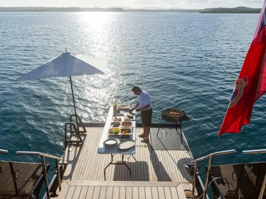 A Chef on ASAHI yacht prepares BBQ lunch at anchor off Fteri Beach, part of your luxury Mediterranean sea yacht charter.