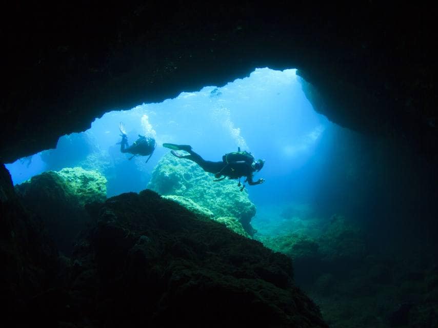 Scuba diving Menorca in turquoise waters, divers enter an underwater cave within a protected marine reserve.