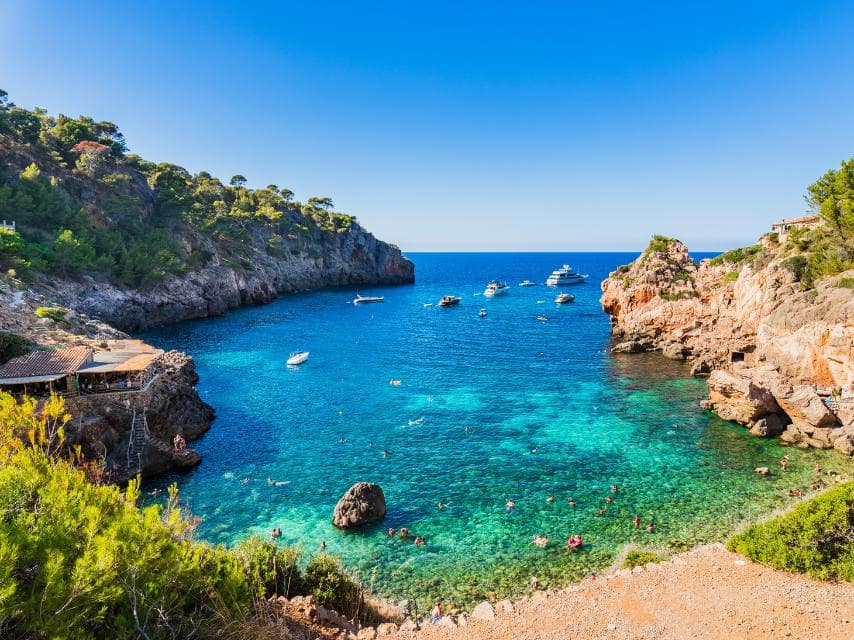Balearic Islands yacht charters anchor in the turquoise waters beside towering cliffs and colorful coral caverns.