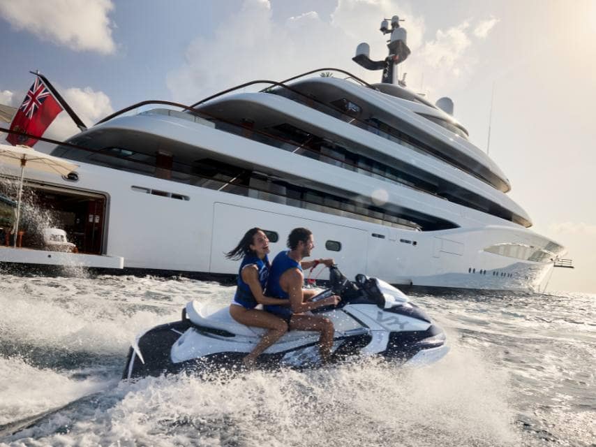 A couple enjoys Jetskis near the SOPHIA yacht, enjoying the water as the yacht’s registration flag flies proudly at the stern.