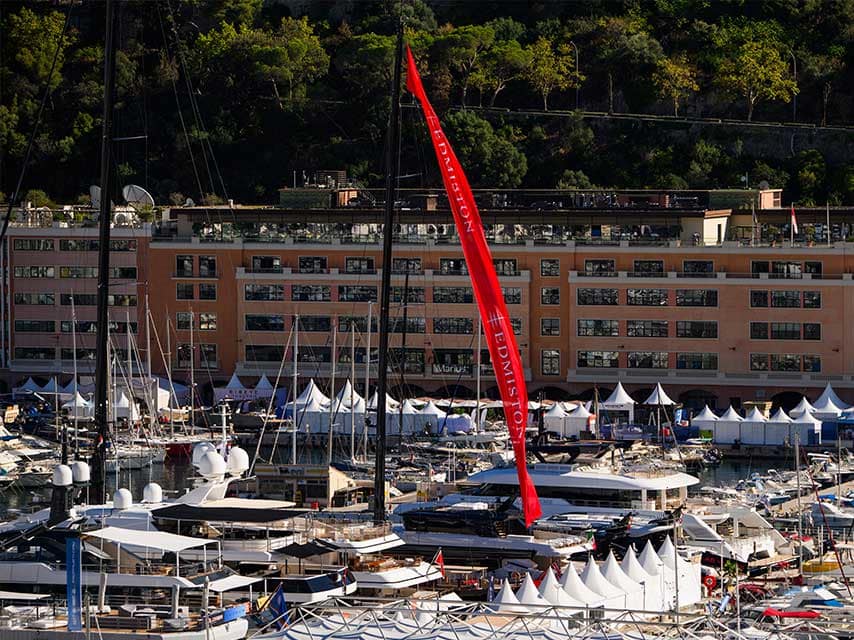 Attend exclusive yacht events at the Monaco Yacht Show with Edmiston, showing their long red banner flying over superyachts in Port Hercule.