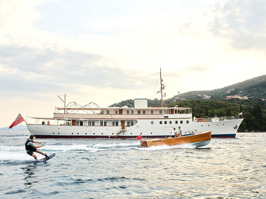 A Corfu yacht charter guest enjoys waterskiing behind the wooden tender of the classic superyacht 'Malahne', anchored nearby.