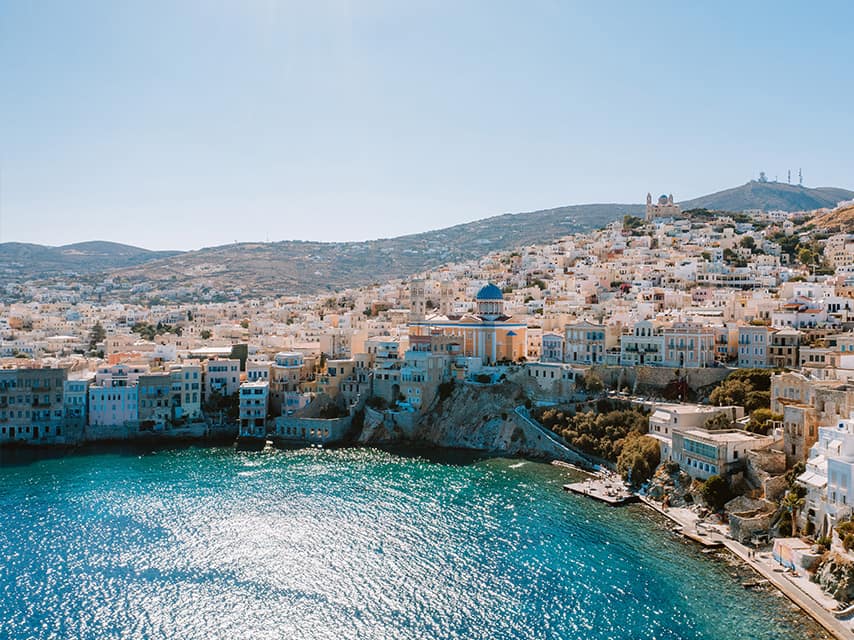 A Cyclades yacht charter reveals Ermoupoli, Syros, with its pastel waterfront and hillside homes by the sea.
